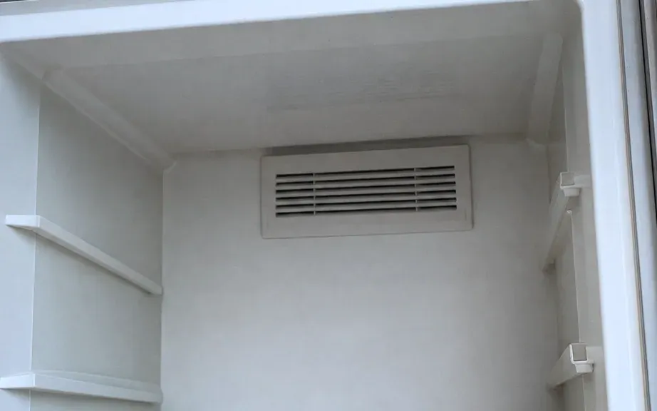 Empty white freezer compartment with a louvered air vent on the back wall and shelf supports on both sides.