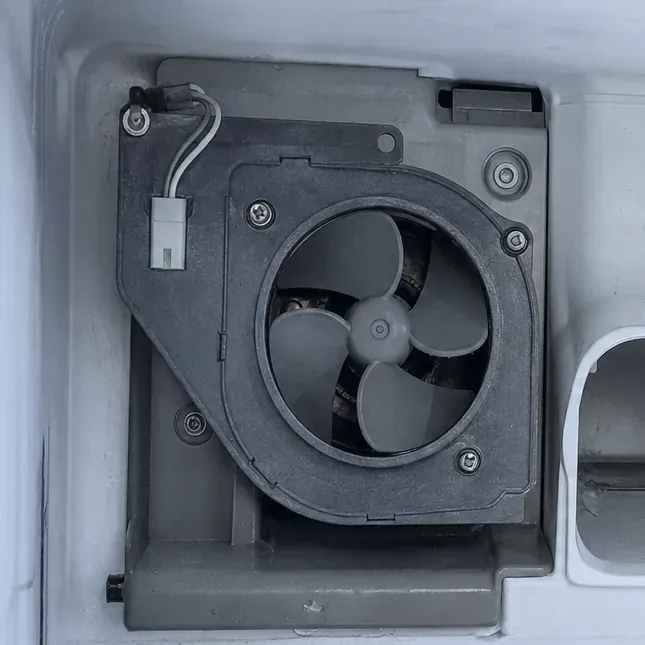 Dark grey fan housing with a three-blade fan mounted on the back wall of a white freezer compartment.
