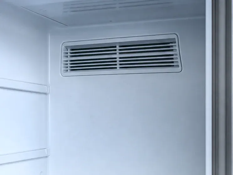 Louvered air vent on the back wall of an empty white compartment, shelf rails along the left side.